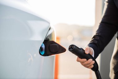 Erkek eliyle elektrik kablosunu açık hava elektrik santralinde beyaz elektrikli arabaya yerleştiriyor. Arka plan bulanık. Bilinçli sahibi olan çevre dostu ulaşım.