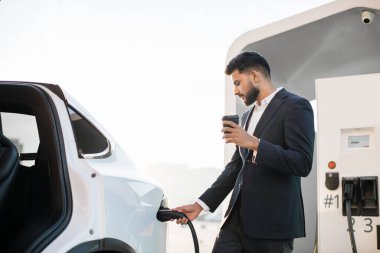 Resmi kıyafetli yakışıklı bir Arap 'ın bir elinde kahveyle lüks elektrikli arabaya fişi takarken görüntüsü. Modern eko taşımacılığın şarj süreci.