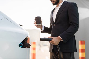 Tarz sahibi tanınmayan işadamlarının yakını elinde elektrik kablosu ve kahve fincanıyla beyaz elektrikli arabanın yanında dikiliyor. Kentsel alandaki EV şarj istasyonu.
