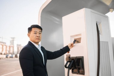 Açık hava istasyonunda elektrikli arabanın parasını öderken kredi kartı kullanan şık siyah takım elbiseli yakışıklı bir adamın portresi. Otomotiv endüstrisinde modern teknoloji ve ulaşılamaz satın alma.