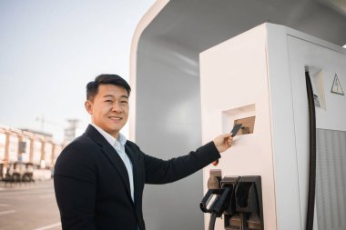 Açık hava istasyonunda elektrikli arabanın parasını öderken kredi kartı kullanan şık siyah takım elbiseli yakışıklı bir adamın portresi. Otomotiv endüstrisinde modern teknoloji ve ulaşılamaz satın alma.