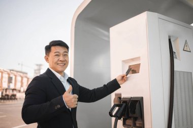 Havalı siyah takım elbiseli yakışıklı bir adamın portresi kredi kartı kullanırken açık hava istasyonunda parmakla gösterilen elektrikli arabanın parasını ödüyor. Otomotiv endüstrisinde modern teknoloji.