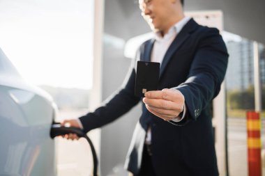 Siyah takım elbiseli pozitif Asyalı adam beyaz elektrikli arabayı şarj ediyor ve açık havada siyah kredi kartı gösteriyor. EV istasyonunda enerji için nakit ödeme.