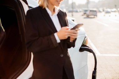 Modern akıllı telefonlu resmi kıyafetler giymiş, dışarıda elektro arabanın yanında duran hoş, beyaz bir kadına yakın durun. Kentsel alanın bulanık arkaplanı.