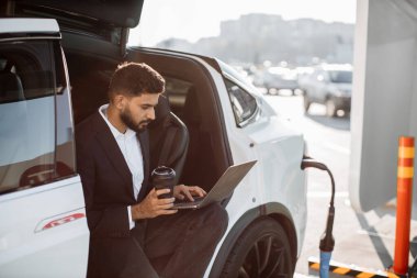 Takım elbiseli, kahve içen ve dizüstü bilgisayarla uğraşan modern elektro arabanın pilini şarj eden bir adam. İnsan kavramı, ulaşım ve teknoloji.