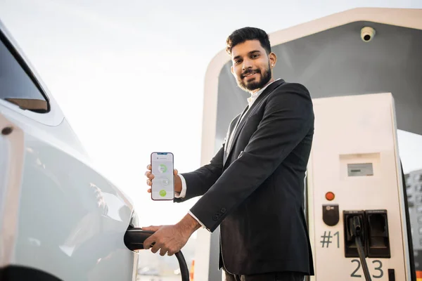 Positive arabian man in business suit charging his electric car at outdoor station and using modern smartphone. Happy owner of eco friendly transport checking battery condition in app.