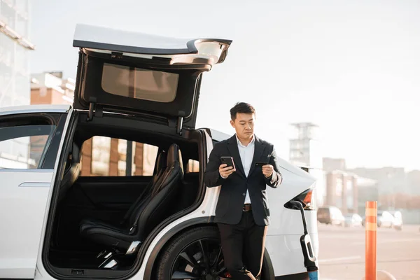 Focused asian man in business suit standing near charging electric car and using smartphone with bank card for paying online. Modern technology for urban lifestyles.