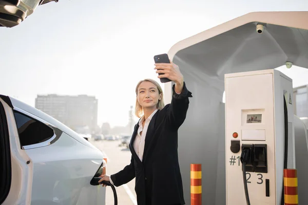 Cheerful caucasian woman in business suit having video call on modern smartphone while charging car at station. Busy modern lifestyles and innovation concept.