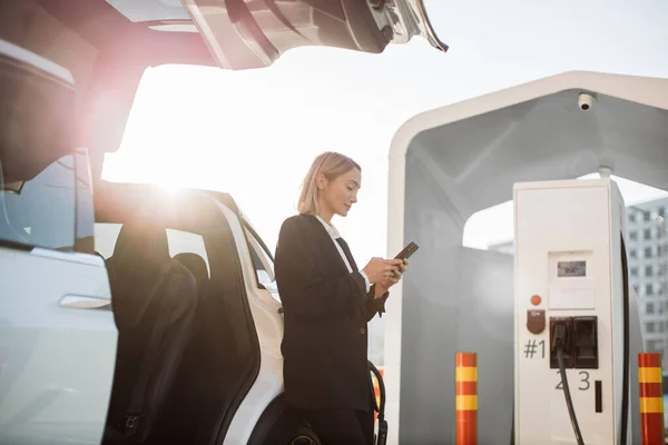 Konzentrierte Geschäftsfrau Mit Smartphone Der Hand Steht Neben Weißem Elektroauto — Stockfoto