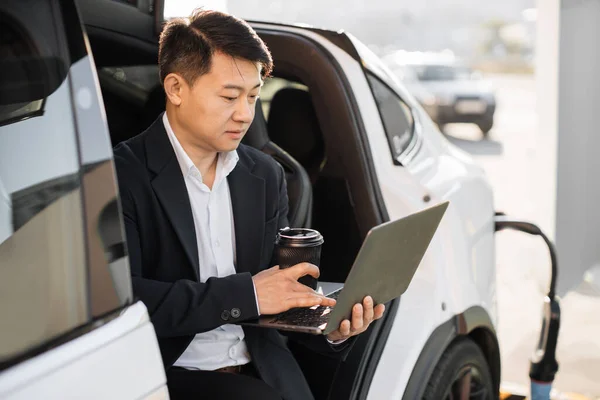 Focused asian man in business suit drinking coffee and working on laptop while charging battery of modern electro car. Concept of people, transport and technology.
