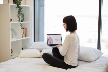 Koyu renk saçlı bir kadının beyaz yatakta oturması ve satılık moda kıyafetlerle web sitesine göz atması. Beyaz kadın kablosuz dizüstü bilgisayar kullanarak% 25 indirimli yeni kıyafet seçiyor..
