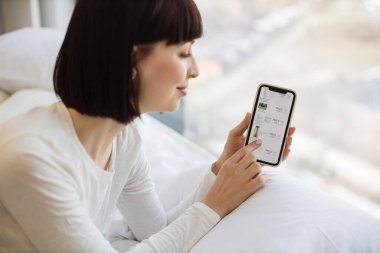 Kahverengi saçlı mutlu çekici bayan internet sitesinde gezinirken profesyonel kozmetik ürünlerini seçiyor. Rahat bir evde online alışveriş yapmak için modern akıllı telefonu kullanan günlük kıyafetli beyaz bir kadın..