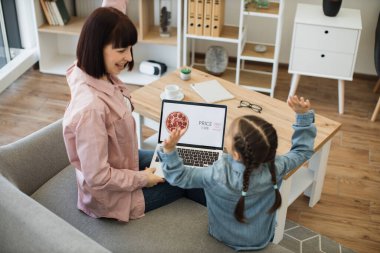 Mutlu genç anne ve tatlı kızı, modern laptop ve neşeli kutlamalarla internetten pizza sipariş ediyorlar. Beyaz kadın ve küçük kız evde yemek pişirmeden eğleniyorlar..