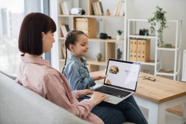 Güzel kızıyla kanepede oturan ve indirimli fast food seçen çekici siyah saçlı kadın. İki kişilik Kafkas ailesi yemek siparişi vermek için modern laptop kullanıyor..