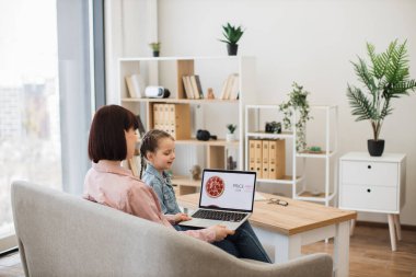 Kafkasyalı siyah saçlı anne ve sevgili kızı rahat oturma odasında dinleniyorlar ve yemek siparişi vermek için modern dizüstü bilgisayar kullanıyorlar. Sıradan giyinen mutlu bayanlar indirimli online pizza seçiyor..