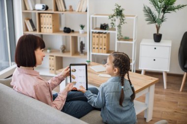 Mutlu anne ve kız kanepede dinleniyor ve elleriyle internetteki abur cubur uygulamasının gösterildiği tablet ekranı işaret ediyorlar. Neşeli bayan müşteriler e-ticaretten faydalanıyor..