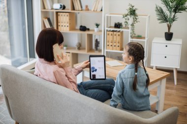 Esmer, beyaz bir kadın telefonda konuşuyor ve yanında küçük bir kız otururken modern bir aleti elinde tutuyor. Genç bayanlar sanal kıyafet mağazasının internet sitesinden günlük kıyafet sipariş ediyorlar..