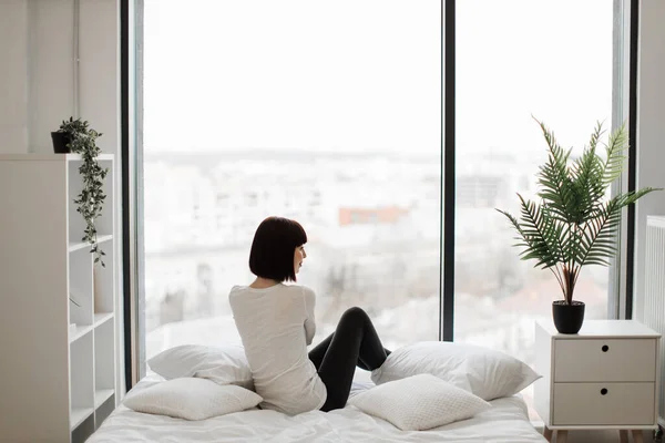 stock image Attractive caucasian woman in domestic outfit resting on white bed with panoramic windows on background. Leisure time at home and lifestyles concept.