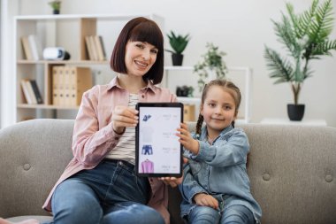 Güzel çocuklu gülümseyen güzel bir bayan elinde tabletle ekranda kıyafet indirimi yapıyor. Çevrimiçi moda mağazalarına göz atarken e-ticaretten hoşlanan mutlu beyaz kadınlar..