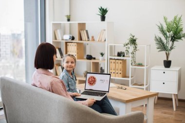 Kafkasyalı siyah saçlı anne ve sevgili kızı rahat oturma odasında dinleniyorlar ve yemek siparişi vermek için modern dizüstü bilgisayar kullanıyorlar. Sıradan giyinen mutlu bayanlar indirimli online pizza seçiyor..
