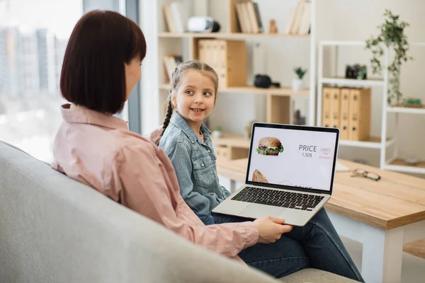 Attractive dark haired woman sitting on couch with her lovely daughter and choosing fast food with discount on website. Caucasian family of two using modern laptop for ordering takeaway lunch.