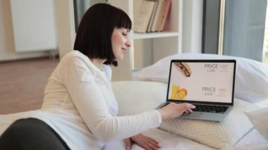 Rahat beyaz kadın rahat bir yatakta kablosuz bilgisayarla uzanıyor ve fast food alışverişi yapıyor. Çeşitli indirimlerle dolu çevrimiçi mağazası olan bilgisayar ekranı.