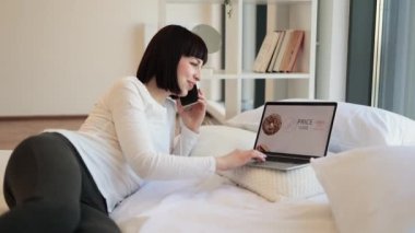 Mutlu kadın esmer cep telefonuyla konuşuyor ve modern dizüstü bilgisayarında yiyeceklerle çevrimiçi alışveriş yapıyor. Hoş bir kadın, günlük giysiler içinde, evde otururken indirimli alışveriş yapıyor..