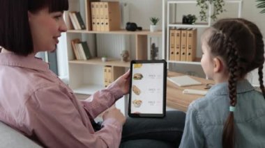 Beyaz dişilerin dijital tablet kullanarak tatlıyı indirimli servis uygulamasında seçmelerini kapatın. Anne ve kızı evde dinlenirken çevrimiçi yemek sipariş etmenin avantajlarından faydalanıyorlar..