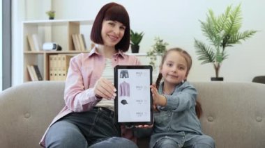 Güzel çocuklu gülümseyen güzel bir bayan elinde tabletle ekranda kıyafet indirimi yapıyor. Çevrimiçi moda mağazalarına göz atarken e-ticaretten hoşlanan mutlu beyaz kadınlar..