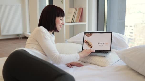 漂亮的年轻女人在舒适的家里很放松 还在网上购物用的是现代笔记本电脑 白种人的黑发在打折期间在市场上订购快餐 专注于屏幕 — 图库视频影像