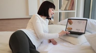 Rahat beyaz kadın rahat bir yatakta kablosuz bilgisayarla uzanıyor ve fast food alışverişi yapıyor. Çeşitli indirimlerle dolu çevrimiçi mağazası olan bilgisayar ekranı.