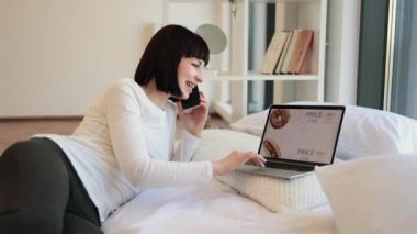 Mutlu kadın esmer cep telefonuyla konuşuyor ve modern dizüstü bilgisayarında yiyeceklerle çevrimiçi alışveriş yapıyor. Hoş bir kadın, günlük giysiler içinde, evde otururken indirimli alışveriş yapıyor..