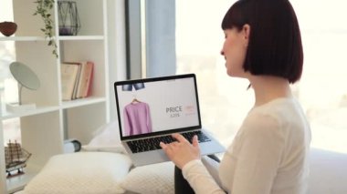 Koyu renk saçlı bir kadının beyaz yatakta oturması ve satılık moda kıyafetlerle web sitesine göz atması. Beyaz kadın kablosuz dizüstü bilgisayar kullanarak% 25 indirimli yeni kıyafet seçiyor..