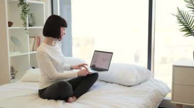 Kahverengi saçlı, beyaz bir kadın, laptopa bakarken mutlu bir şekilde el ele tutuşuyor. Kadın müşteri indirimli kıyafet alırken çok mutlu oluyor..
