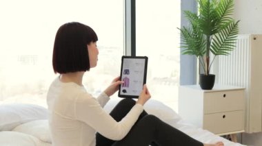 Koyu renk saçlı beyaz kadının internet sitesinde dijital tablet kullanarak satışta olan modaya uygun kıyafetlerini çekerken yan görüntüsü. Sıradan giyinmiş bir bayan evde otururken alışverişin keyfini çıkarıyor..