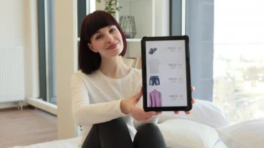 Siyah saçlı, beyaz bir kadının internet sitesinde dijital tablet kullanarak satışta olan modaya uygun kıyafetlere bakması. Sıradan giyinmiş bir bayan evde otururken alışverişin keyfini çıkarıyor..