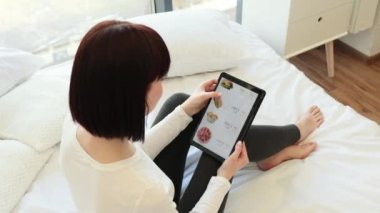 Çekici beyaz kadın, gündelik giysiler içinde elinde dijital masa ile yatakta oturuyor ve internetten fast food satıyor. Market, alışveriş ve alet edevat kavramı.