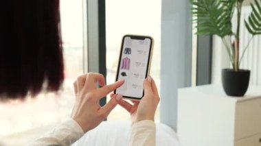 Rahat giyimli hoş beyaz kadın evde dinlenirken internetten yeni moda kıyafetler sipariş etmek için akıllı telefon kullanıyor. Modern aletlerin yardımıyla satış sezonunda alışveriş..