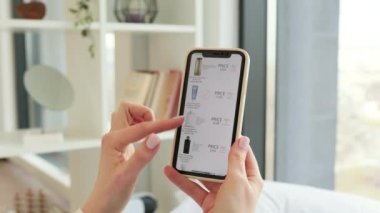 İnternet sitesinde gezinirken profesyonel kozmetik ürünlerini seçen bir kadının elleri. Kafkasyalı bir kadın, konforlu evde online alışveriş için modern akıllı telefonu kullanıyor..