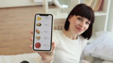 Kafkasyalı güzel bir kadının portresi. Yatakta oturuyor ve modern akıllı telefonu elinde tutuyor. Seyyar ekrana odaklanın, ekommerce çevrimiçi dükkan indirimli yiyecek satıyor..
