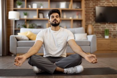 Spor giyimli rahat Hintli adam rahat oturma odasında Lotus pozunda meditasyon yapıyor. Genç sakallı adam Mudra jesti yapıyor ve egzersiz sırasında sakince nefes alıyor..