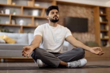 Aktif giyimli yakışıklı sakallı bir Arap gözlerini kapatıyor ve rahat oturma odasındaki yoga minderinde meditasyon yapıyor. Çakra, dzen ve vücut sağlığı kavramı.