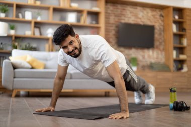 Çekici Arap adam oturma odasındaki yoga minderinin üzerinde tahta pozisyonunda duruyor. Spor kıyafetli genç adam kameraya bakıp gülümsüyor ve düz ellerle dengede duruyor..