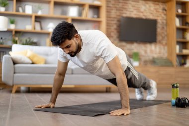 Modern apartman dairesinde egzersiz yaparken spor kıyafetleri giyen güçlü bir Arap adam tahta pozisyonu alıyor. Karın kasları için yoga minderi kullanan odaklanmış genç bir erkek. Dayanıklılık ve güç kavramı.