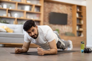 Spor elbiseli sporcu Arap adam evde antrenman yaparken karın kaslarını tahta pozisyonunda çalıştırıyor. Yakışıklı sakallı adam sabahları vücut egzersizi yapıyor..