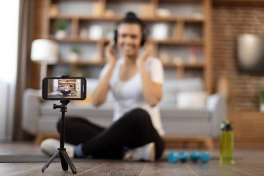 Sağlıklı, çok ırklı, bağdaş kurmuş bir kadın kulaklıkla otururken çevrimiçi paylaşım için fitness eğitiminin video içeriğini kaydediyor. Ekranda spor vlog oynatıcısı olan akıllı telefona odaklan.