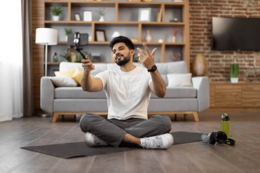 Ayakları çapraz oturan aktif Arap erkek geniş bir odada tripodla cep telefonuyla kişisel antrenman yapıyor. Kendine güvenen fitness blogcusu kamera kullanarak sabah sporunu canlı yayınlıyor.