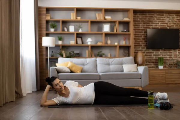Multiethnic charming woman dressed in sport clothes lying on yoga mat at living room and lifting legs up. Concept of body care, healthy lifestyles and regular workout.