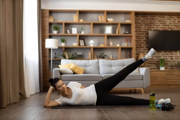 Multiethnic charming woman dressed in sport clothes lying on yoga mat at living room and lifting legs up. Concept of body care, healthy lifestyles and regular workout.