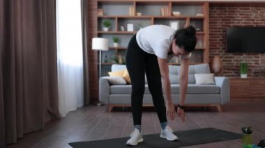 Spor kıyafetleri giymiş çok ırklı bir kadın sıcak ve modern bir dairede sabah egzersizinden önce ısınıyor. Egzersiz, öz disiplin ve vücut bakımı kavramı.